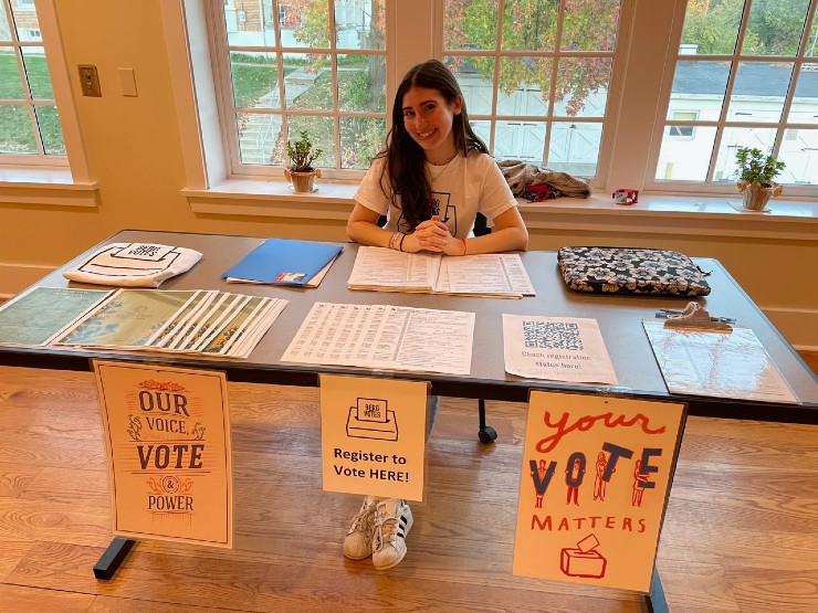 A college student sits behind a table full of information about registering to vote.